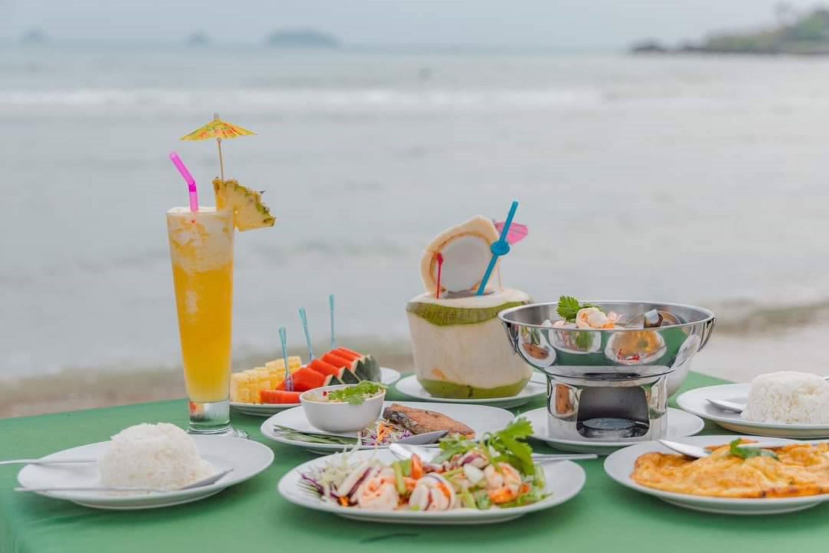 Ein Tisch ist reichhaltig mit Speisen gedeckt, darunter Reis, ein Salat und ein Gericht mit Meeresfrüchten, mit Blick auf das Meer im Hintergrund.