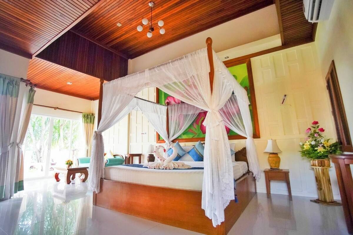 A room with a four-poster bed, a wooden ceiling, and a large window with curtains.