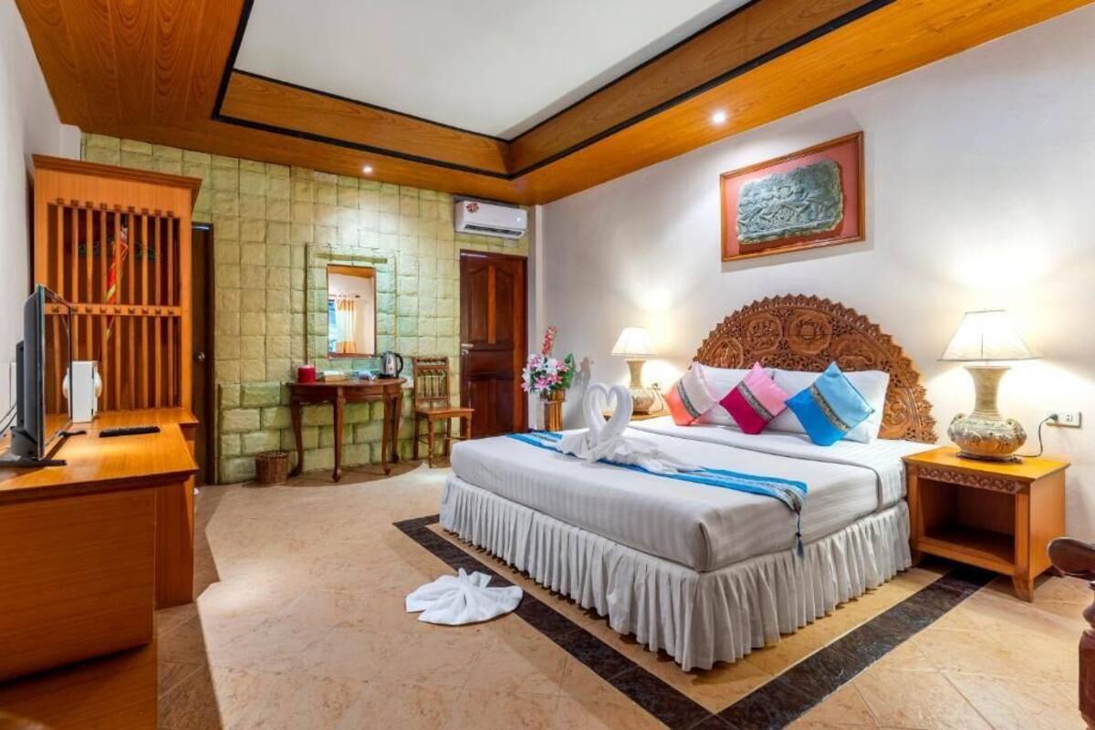 A hotel room with a large bed, wooden headboard, two bedside tables with lamps, a desk with a computer, a stone wall, and a wooden cabinet.