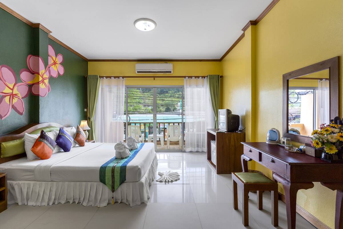 A hotel room with a bed, a desk, a chair, a dresser, and a balcony with a view of greenery.