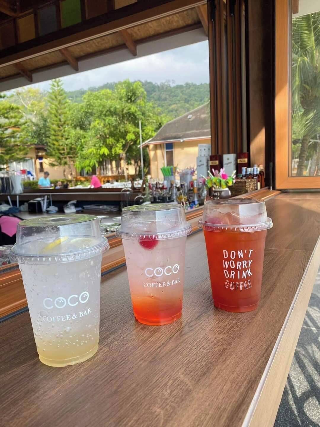 Three beverage cups on a wooden table with a view of a lush green landscape and a building in the background.