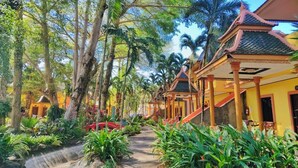 A pathway lined with tropical plants and palm trees, leading to traditional-style buildings with red roofs and yellow walls. - Coconut Beach Resort (Ko Chang)