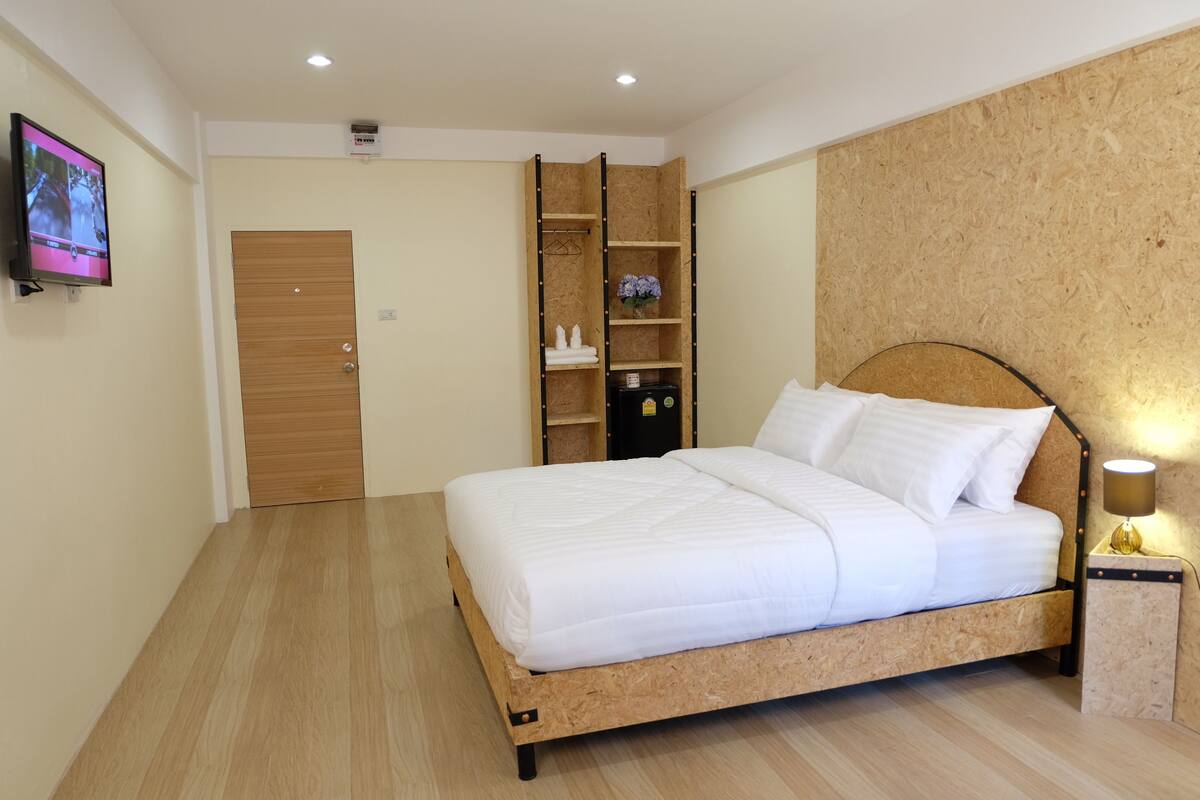 A neatly arranged bedroom with a bed, a wooden wardrobe, a shelf unit, and a television mounted on the wall.