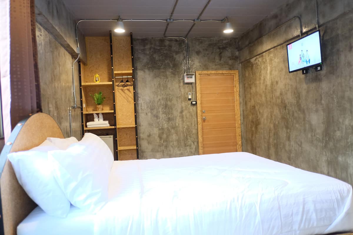 A bedroom with a bed, a wooden wardrobe, a shelf with a plant, and a television on the wall.