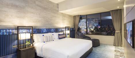 Habitación de hotel moderna con una cama grande, una mesita de noche con lámpara, una silla y vistas nocturnas de la ciudad.