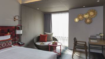 A hotel room with a bed, a seating area with a chair and a small table, a desk with a chair, and a large window with curtains.