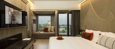 A modern hotel room with a large bed, a flat-screen TV mounted on the wall, a sofa, and a balcony with a city view.