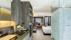 A modern hotel room with a large mirror, a vanity with a sink, a bed, a sofa, and a view of a cityscape.