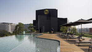 A rooftop infinity pool with lounge chairs and umbrellas, a modern building with a distinctive logo, and a cityscape in the background.