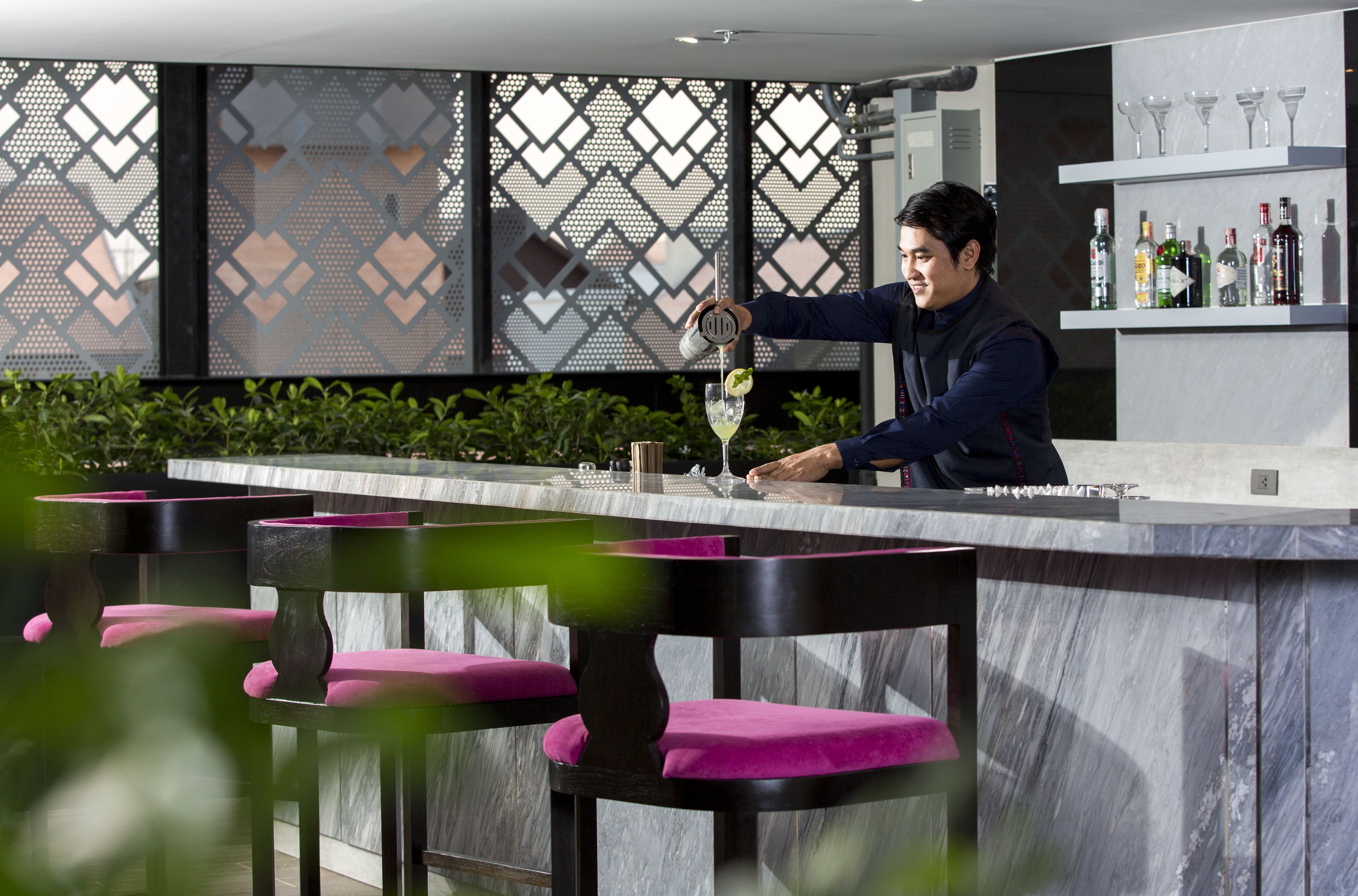 Una zona de bar con encimera de mármol, taburetes negros con cojines rosa y una pared decorativa con motivo geométrico.