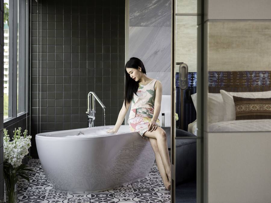 A woman sitting in a freestanding bathtub in a modern bathroom with dark tiles and patterned flooring.