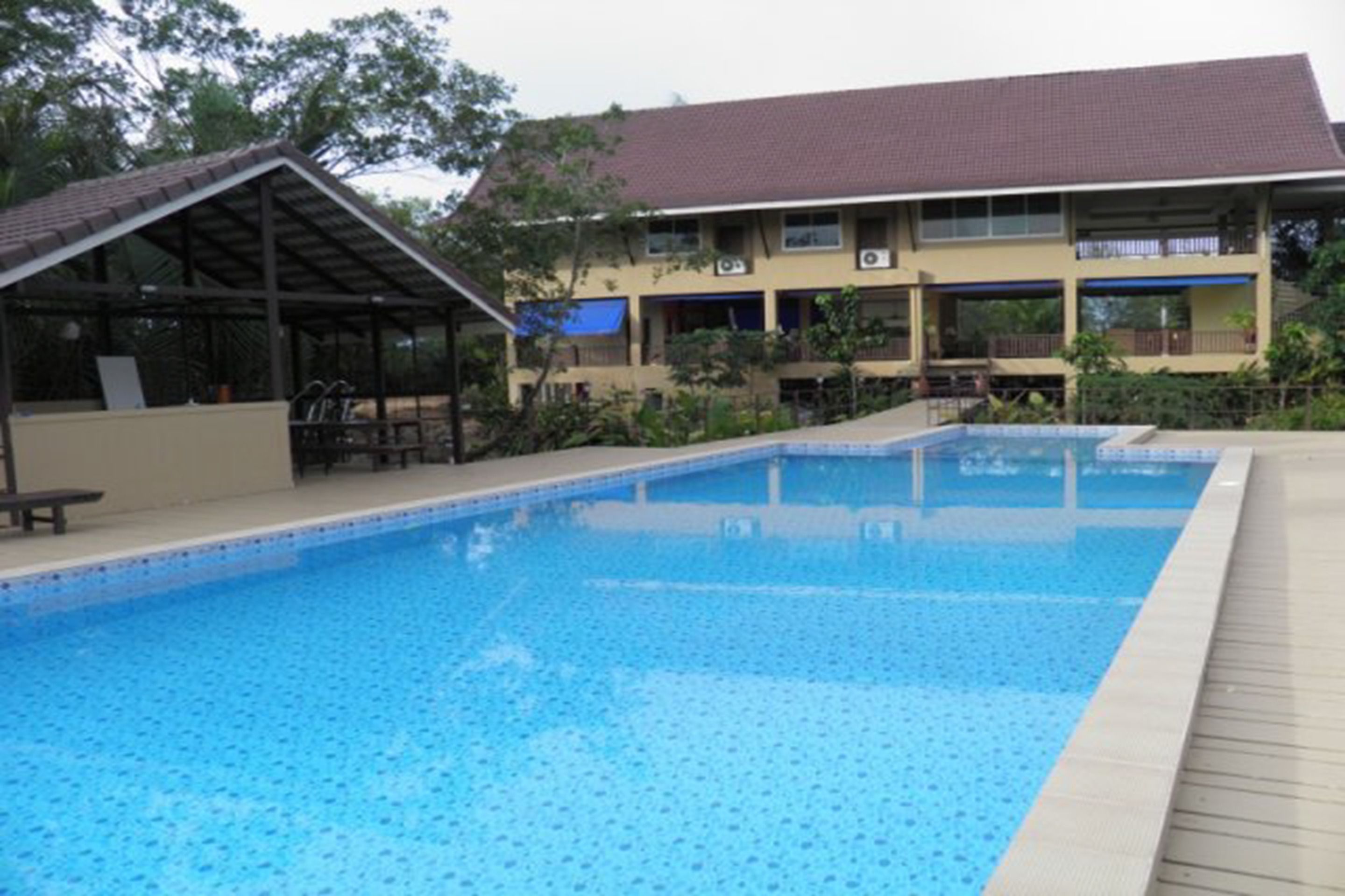 A swimming pool with a tiled deck, a covered seating area, and a building with multiple balconies in the background.