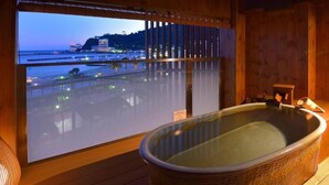 A room with a large bathtub and a view of a beach and cityscape through a window. - Atami Seaside Spa & Resort (Atami)