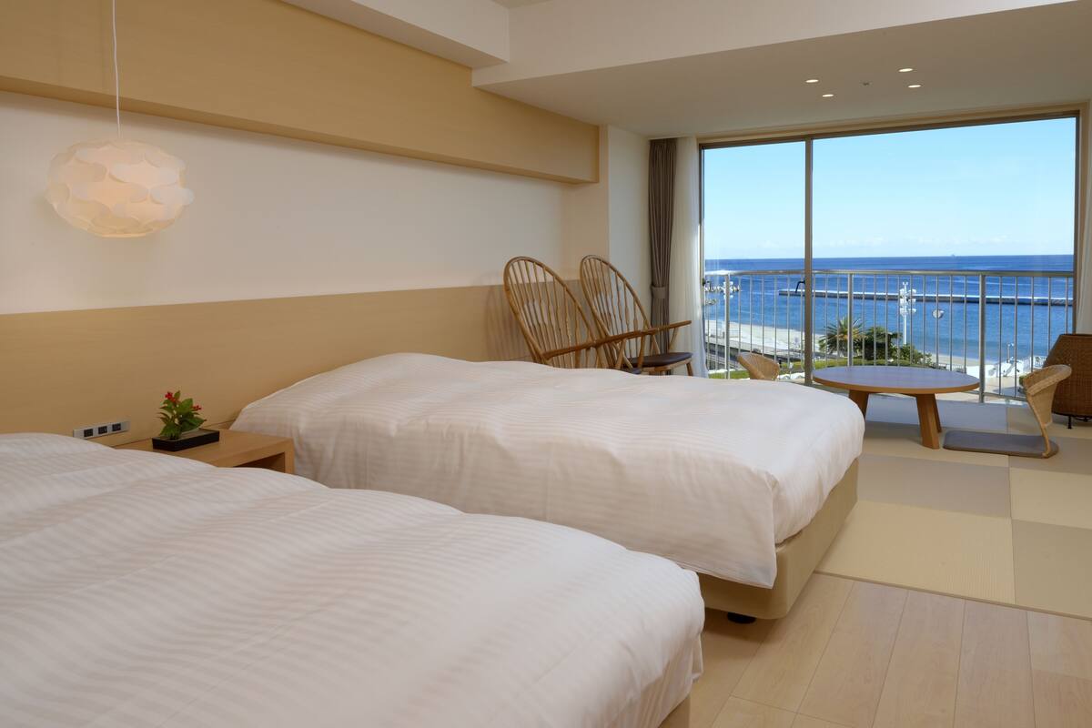 A hotel room with two beds, a balcony with a view of the sea, and a small table.