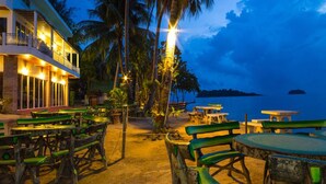 Un restaurante frente al mar con mesas al aire libre, palmeras y vista al océano al atardecer.