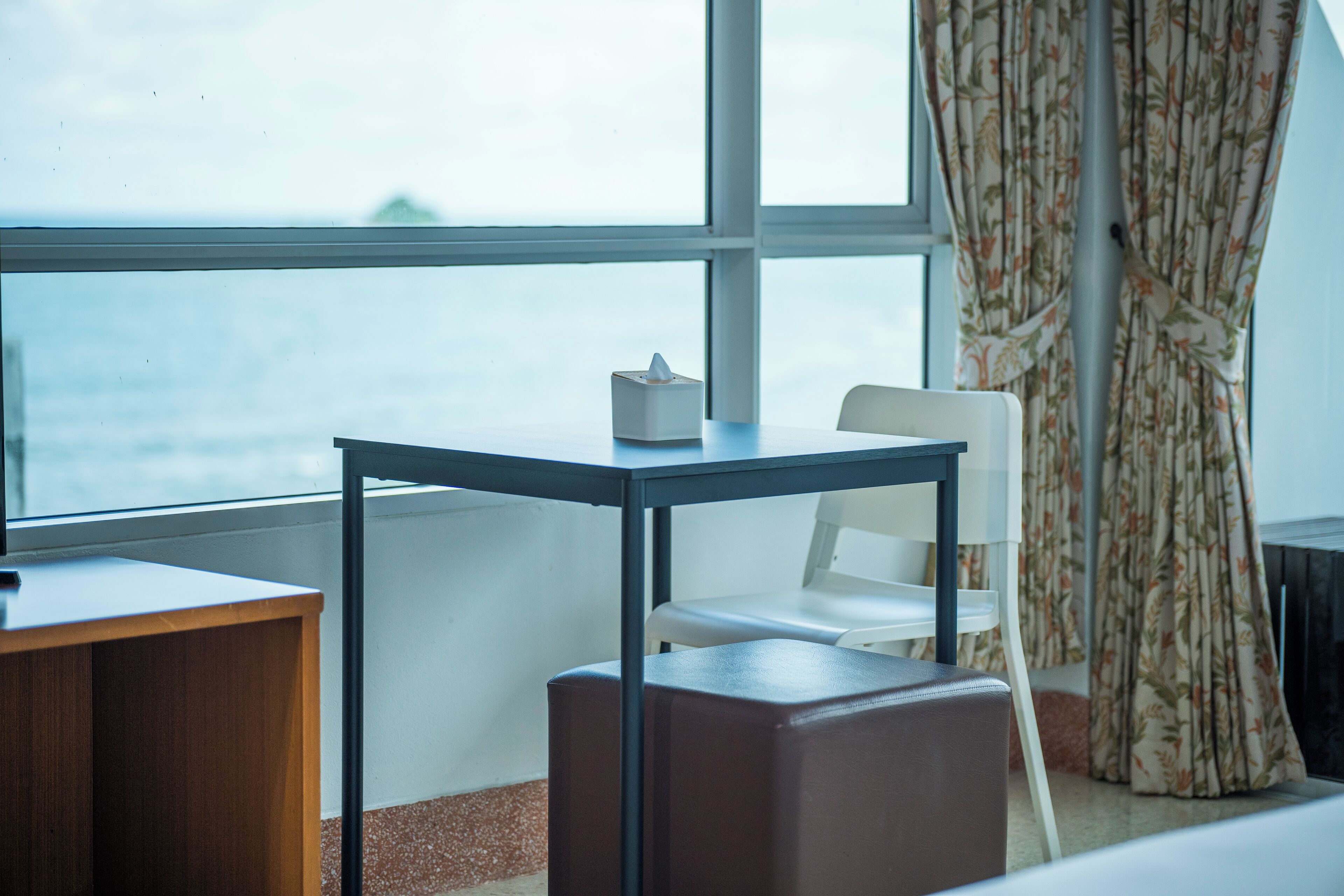 A room with a view of the sea, a table, a chair, and a small stool.