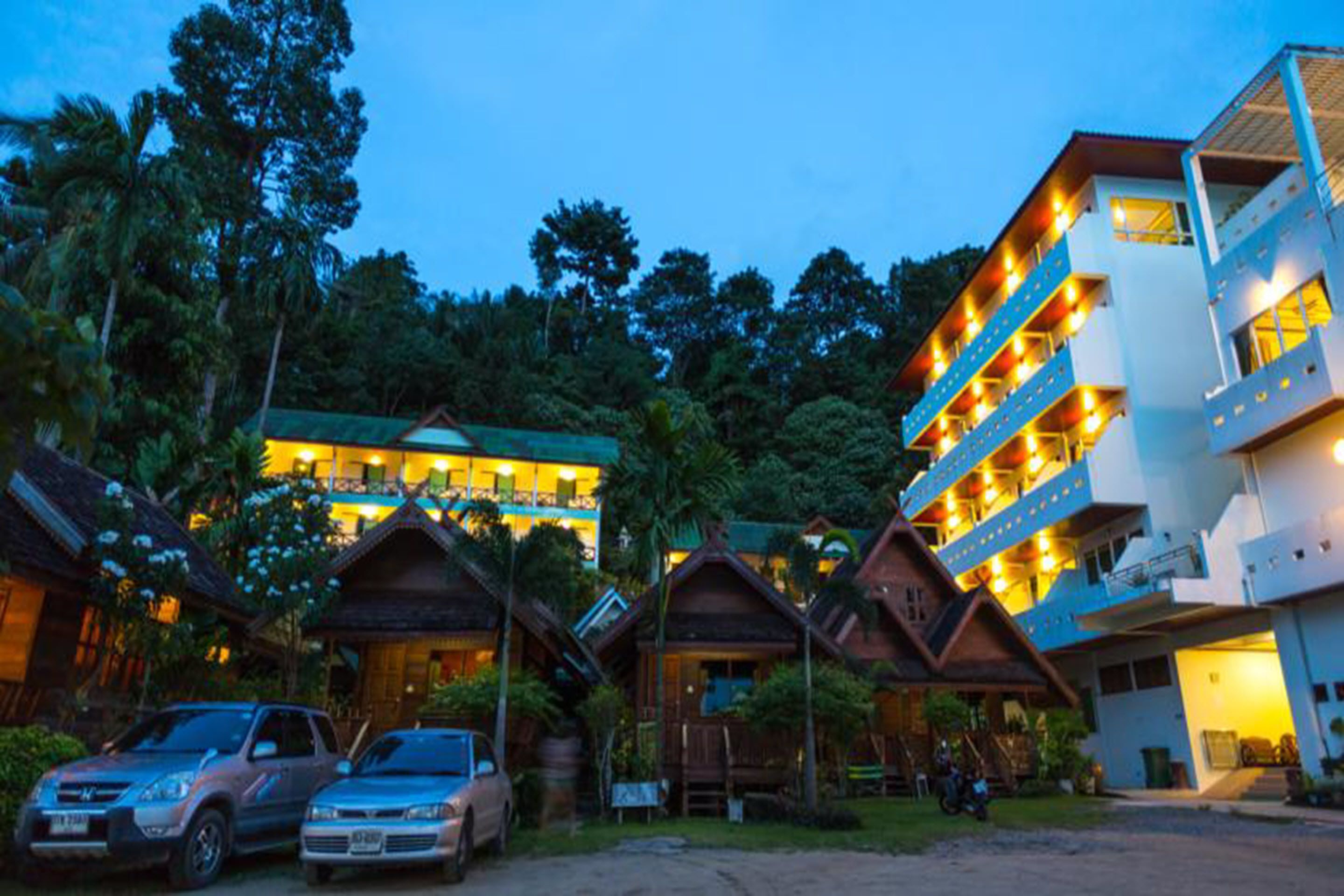 A hotel with multiple-story buildings, a parking area with cars, and lush greenery in the background.