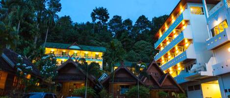 A hotel with multiple-story buildings, a parking area with cars, and lush greenery in the background.