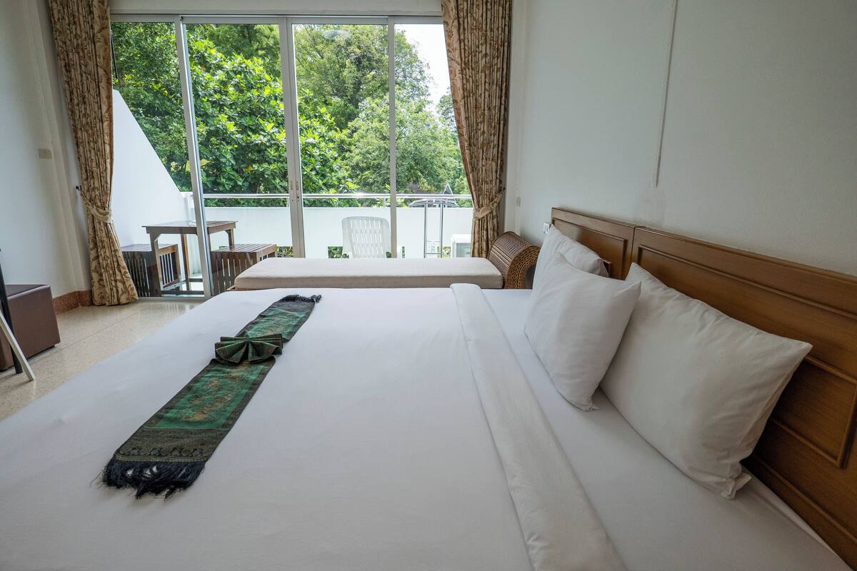 Habitación de hotel con una cama grande, vistas a la vegetación por la ventana y un balcón con mesa y sillas.