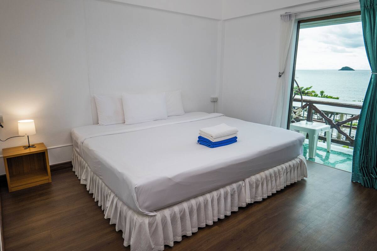 Habitación de hotel con una cama grande, una mesita de noche y vistas al mar a través de una ventana.