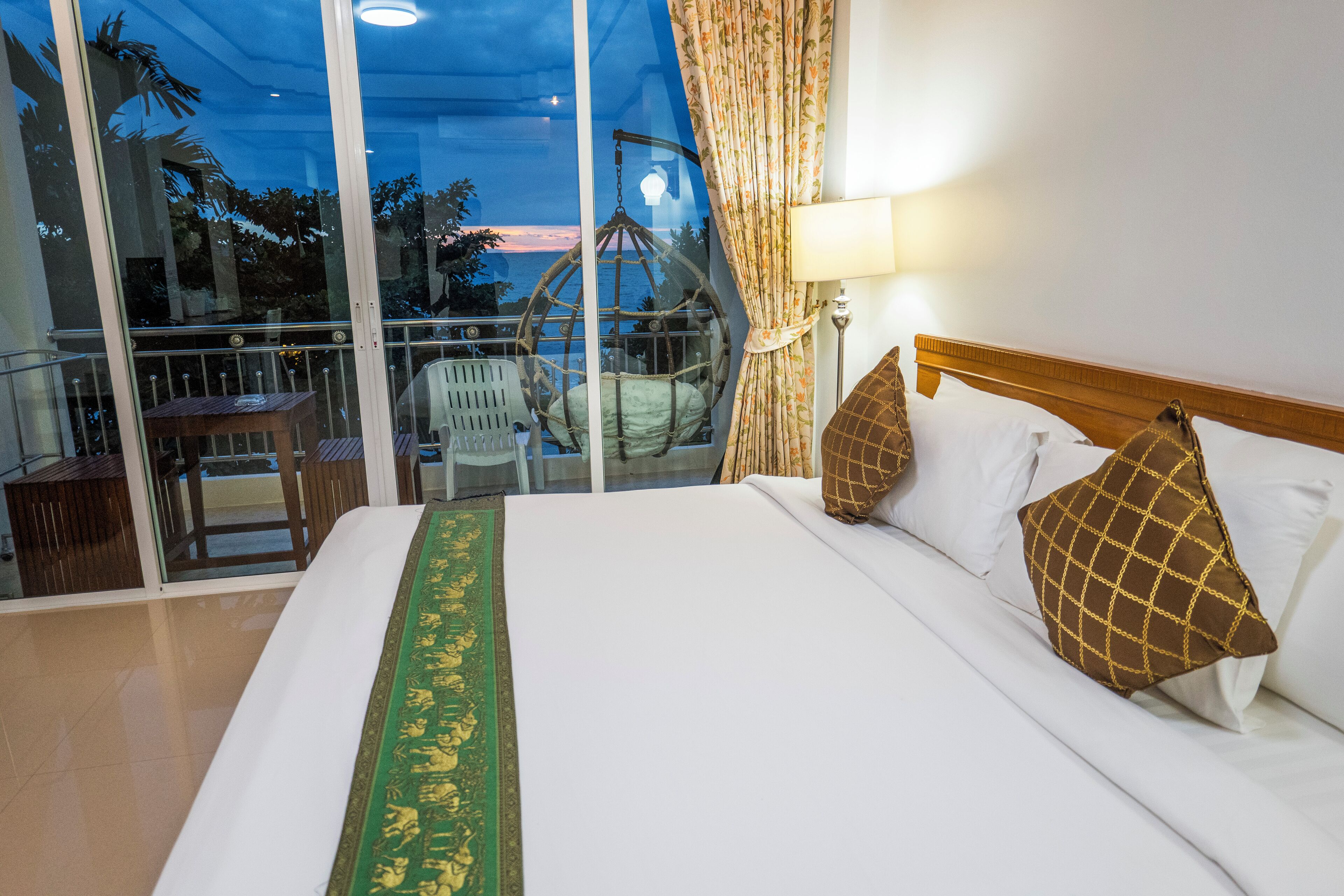 A hotel room with a large bed, a view of the ocean through floor-to-ceiling windows, and a balcony with a hanging chair.
