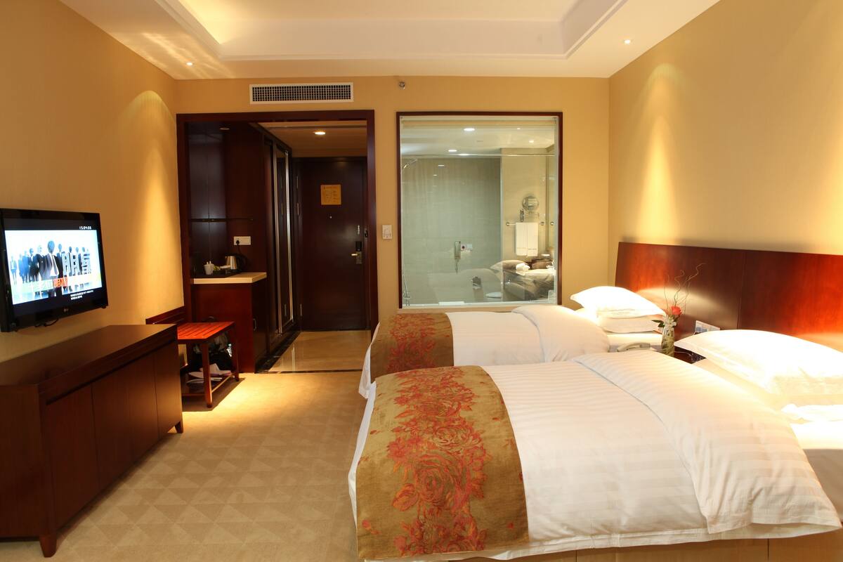 Habitación de hotel con dos camas, televisión, escritorio y baño visible a través de una puerta abierta.