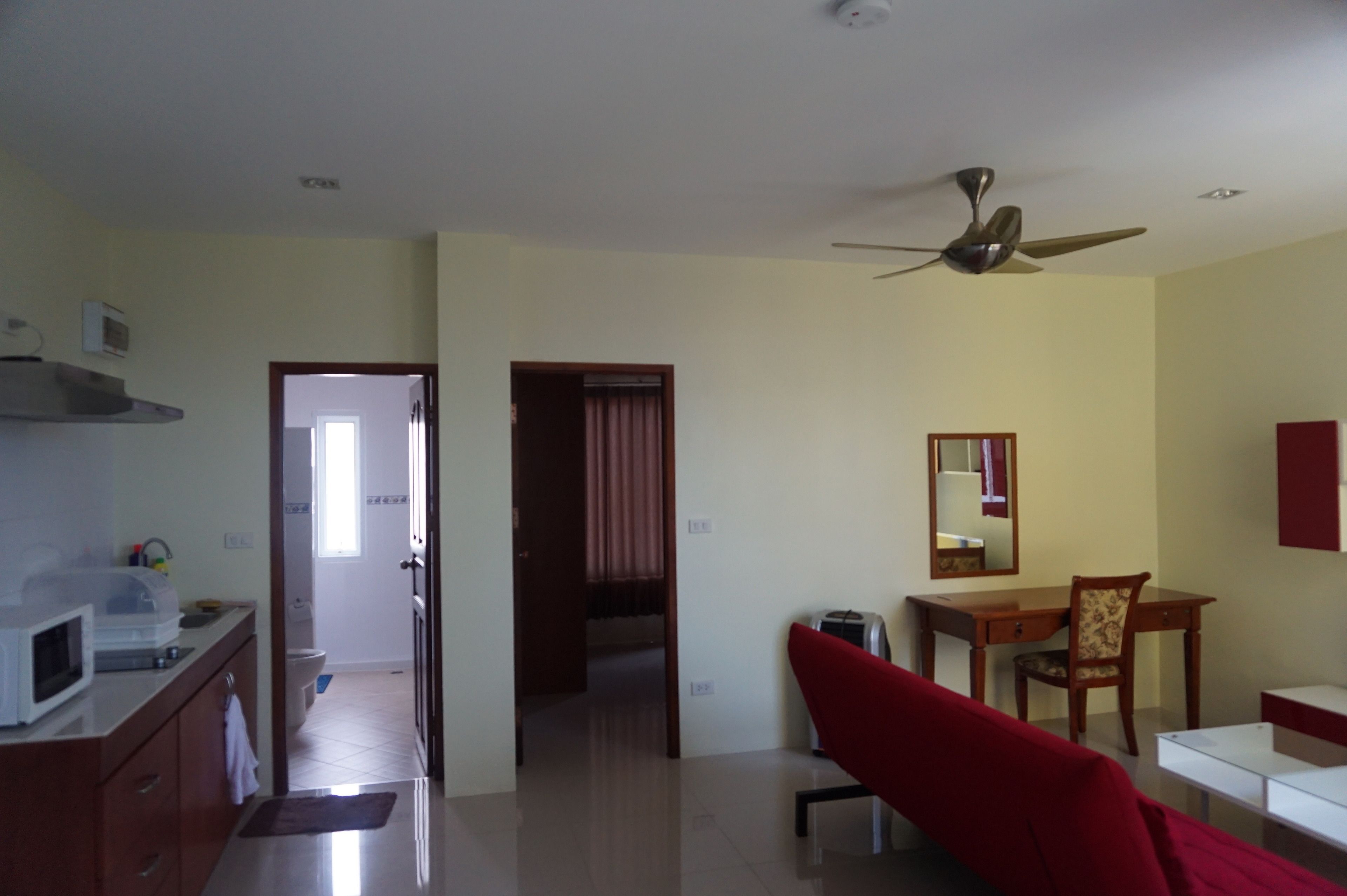 A room with a kitchenette, a dining area with a chair and table, a ceiling fan, and a door leading to another room.