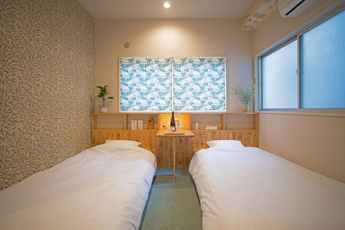 A hotel room with two single beds, a wooden desk with a small table lamp, and a window with a floral pattern.