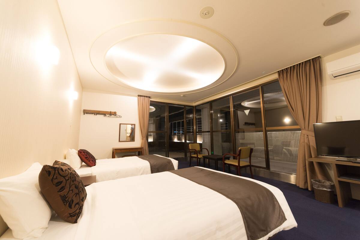 Une chambre d’hôtel avec deux lits, un bureau avec un ordinateur, une chaise et une grande fenêtre avec des rideaux.