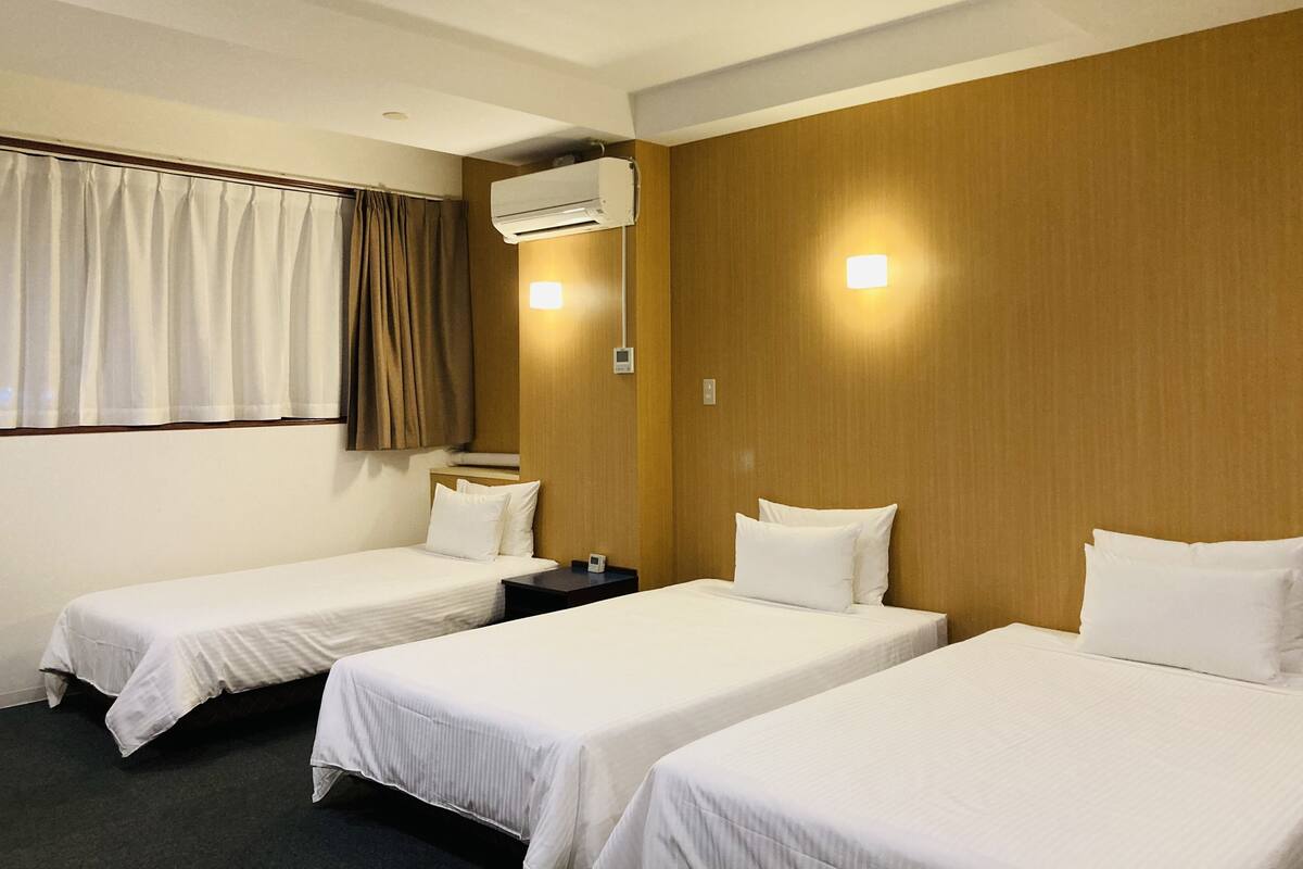 Une chambre d’hôtel avec deux lits simples, une fenêtre avec des rideaux, un appareil de climatisation et une table de chevet avec une lampe.