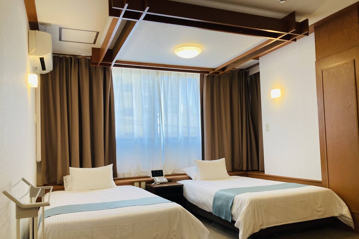 Une chambre d’hôtel avec deux lits, une table de chevet avec un téléphone, une fenêtre avec des rideaux et un luminaire au plafond.