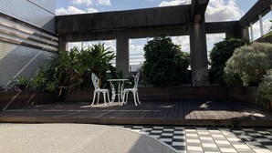 Un espace extérieur aménagé avec des sièges, doté d’un sol à carreaux, entouré de plantes en pot et de chaises blanches.