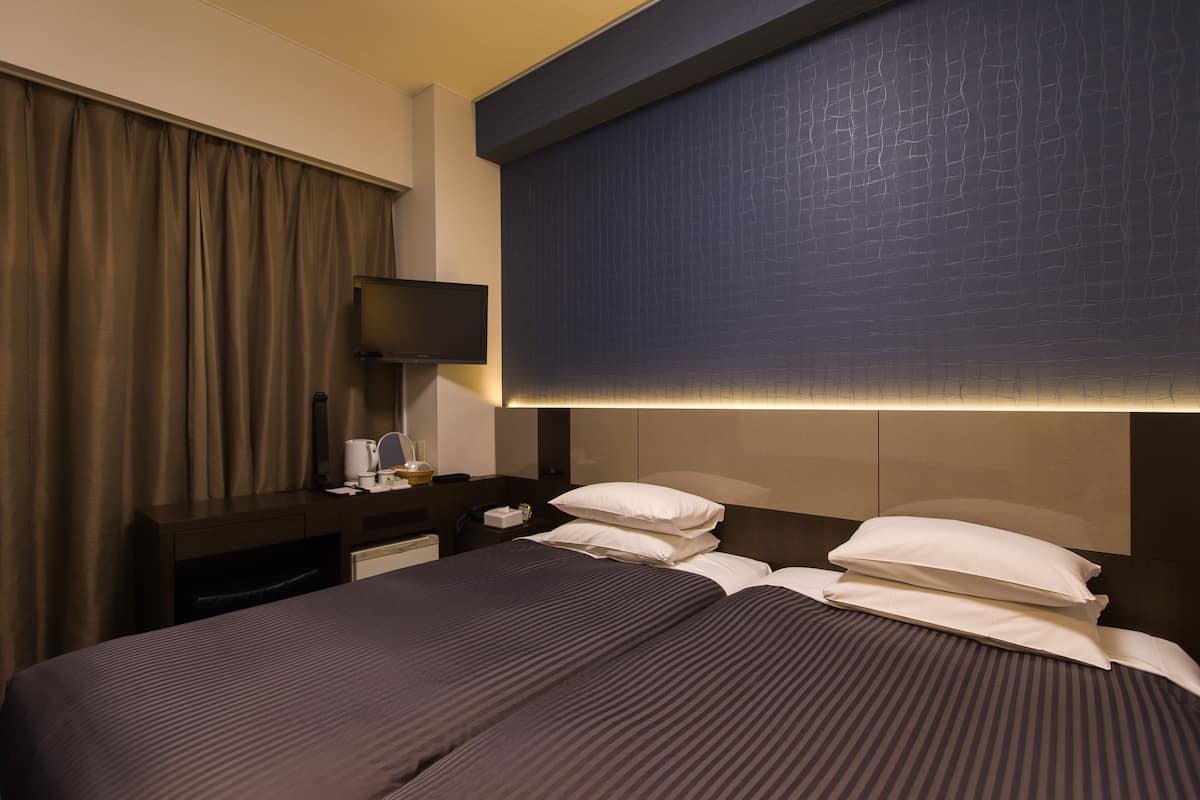 A hotel room with a large bed, a flat-screen TV mounted on the wall, a bedside table with a lamp, and a curtain.