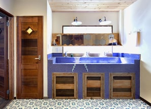 Un baño con puerta de madera, lavamanos con azulejos azules y tocador de madera con espejo arriba.