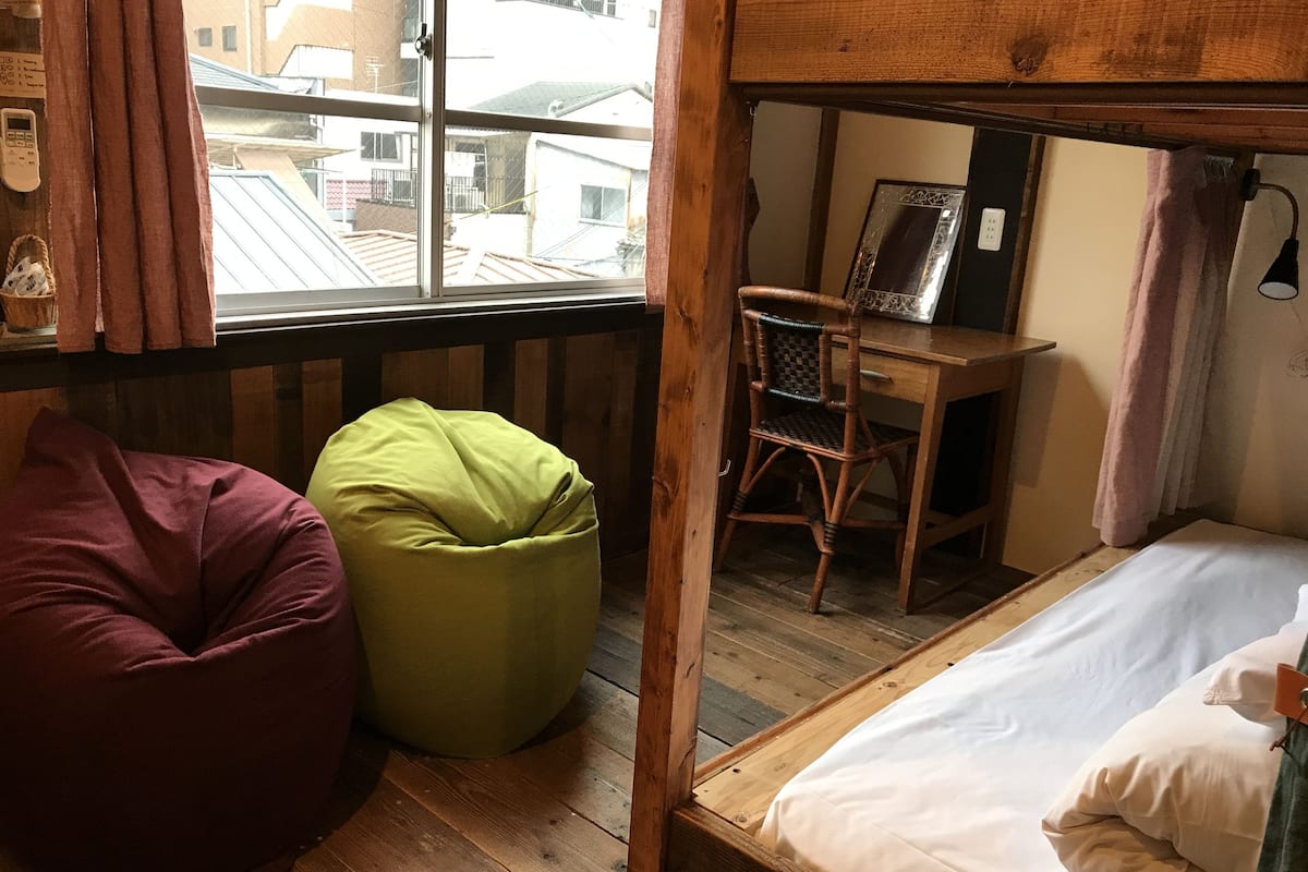 A small, cozy room with a bunk bed, a desk, and a chair. There is a window with a view of buildings outside.