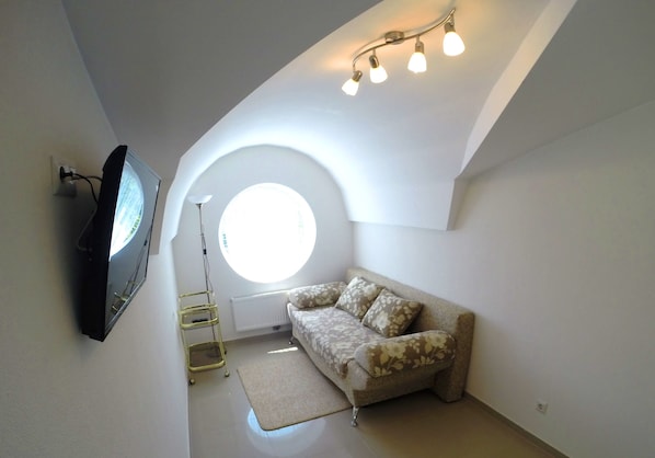A small, well-lit room with a round window, a sofa, a TV mounted on the wall, and a ceiling light fixture.