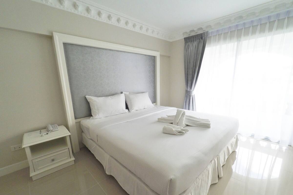 Un grand lit avec du linge de lit blanc, une table de chevet avec un téléphone et une fenêtre avec des rideaux.