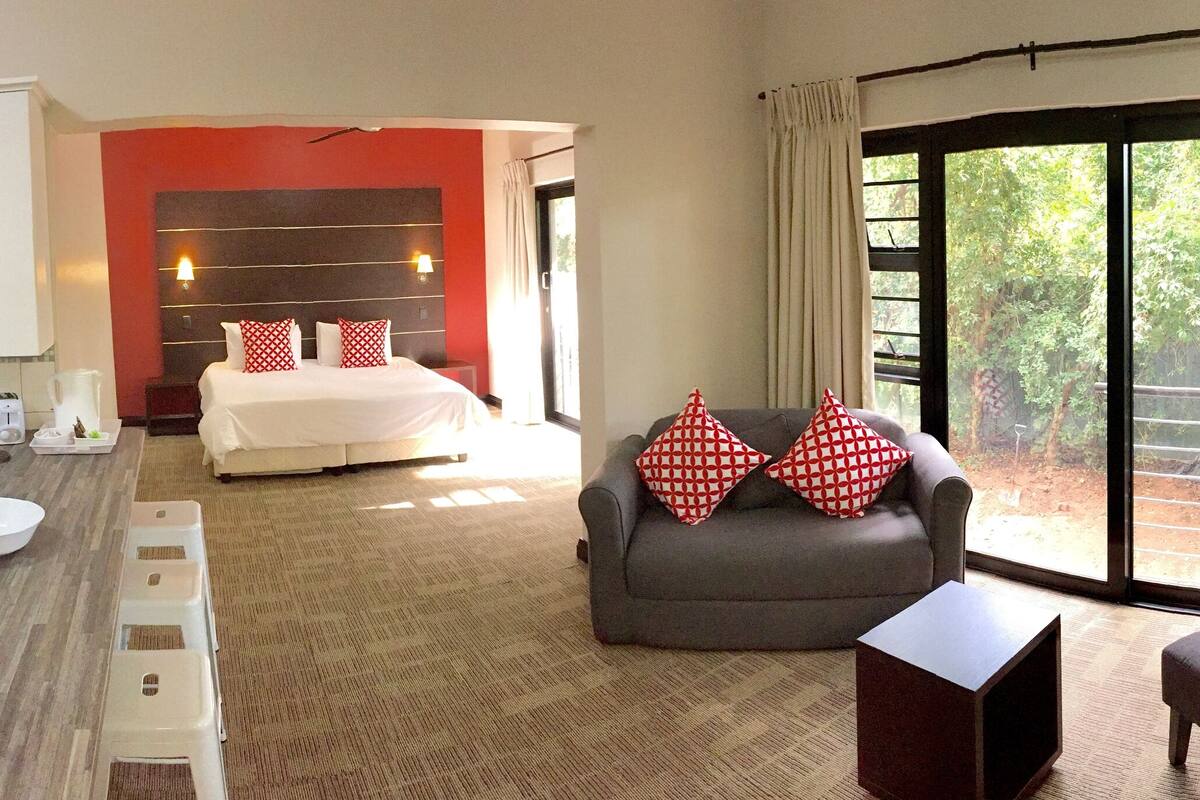 A hotel room with a kitchenette, a bed with red and white patterned pillows, a grey sofa, and a large window with a view of greenery.