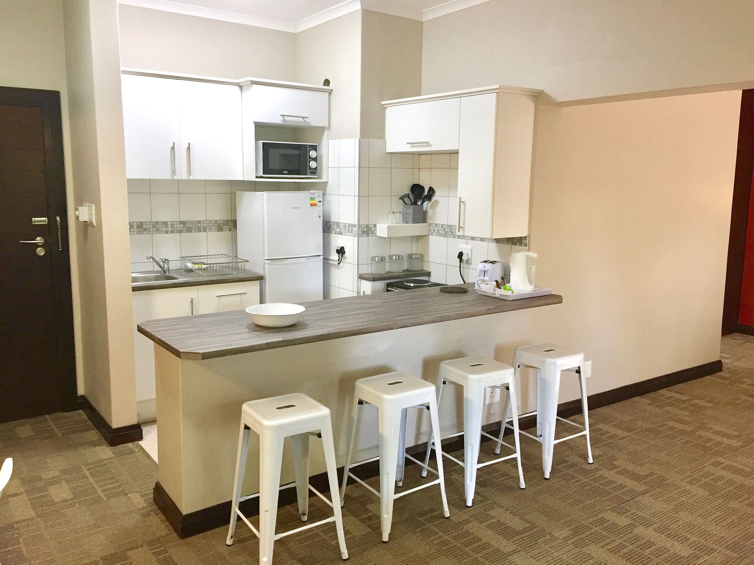 A compact kitchen with white cabinets, a microwave, a refrigerator, a sink, and a bar stool seating area.