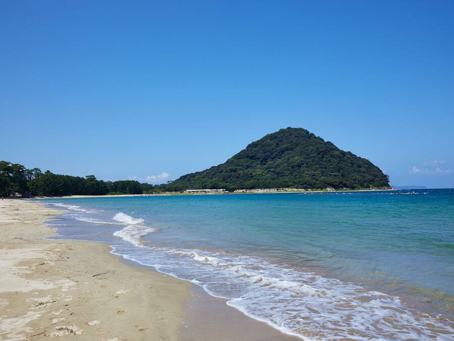 A beach with clear turquoise water, a sandy shore, and a forested hill in the background.