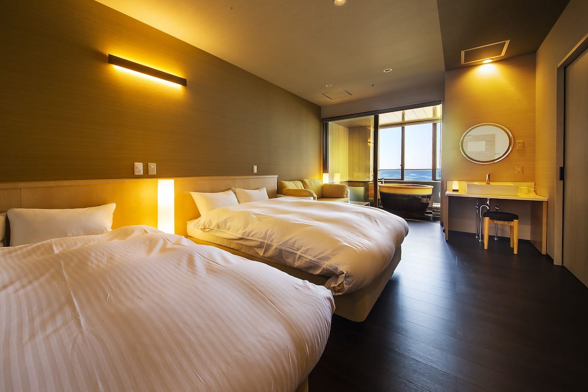 Ein Hotelzimmer mit zwei Betten, einem Schreibtisch mit Stuhl, einem großen Fenster und einem Badezimmer mit Spiegel.