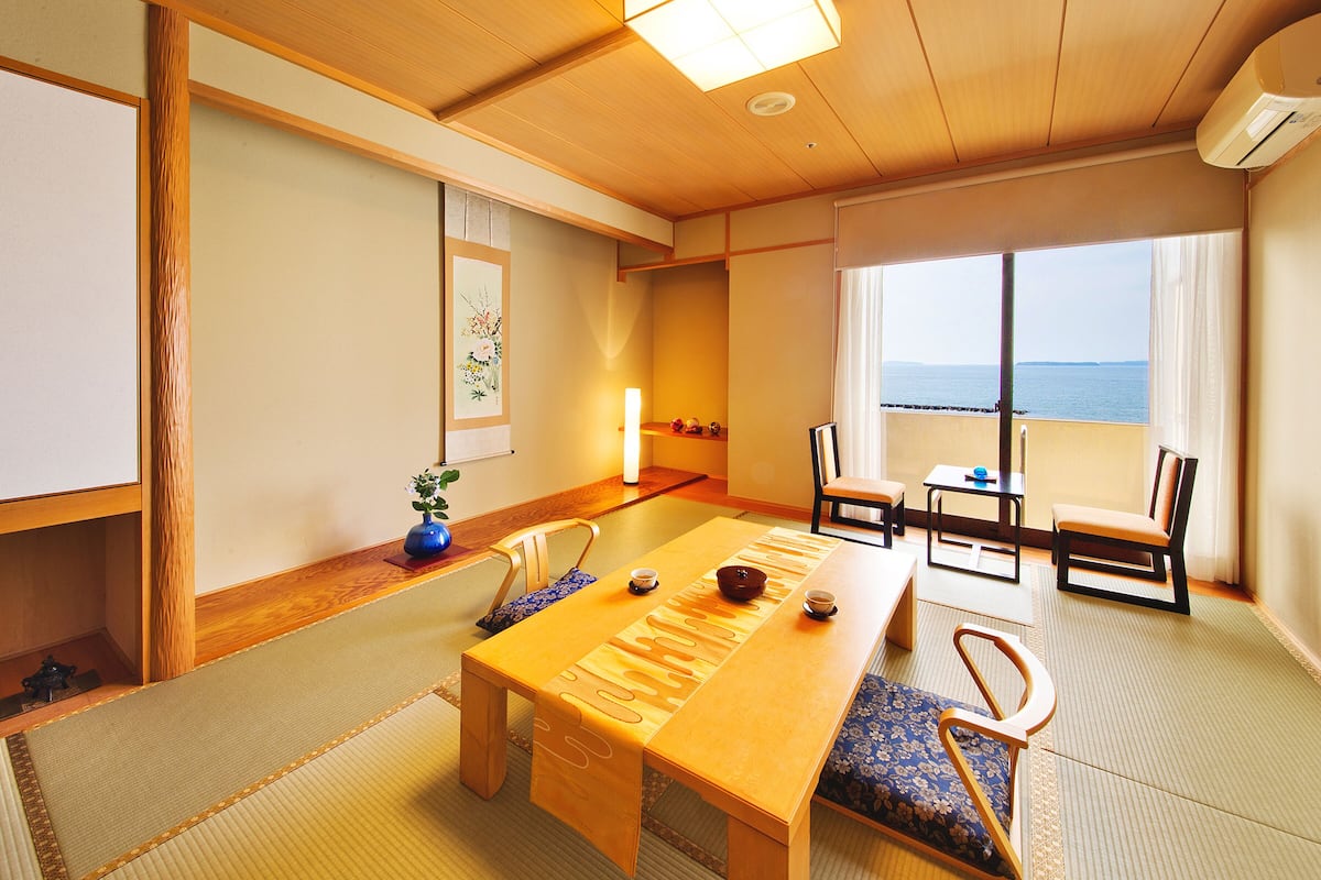 Ein traditioneller japanischer Raum mit Tatami-Böden, einem niedrigen Holztisch und Blick auf das Meer durch große Fenster.