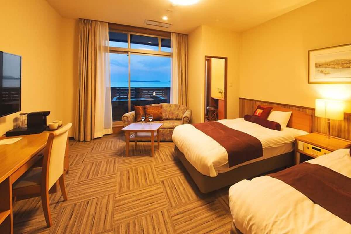 Ein Hotelzimmer mit zwei Betten, einem Schreibtisch, einem Sessel, einem Tisch und einem großen Fenster mit Blick.