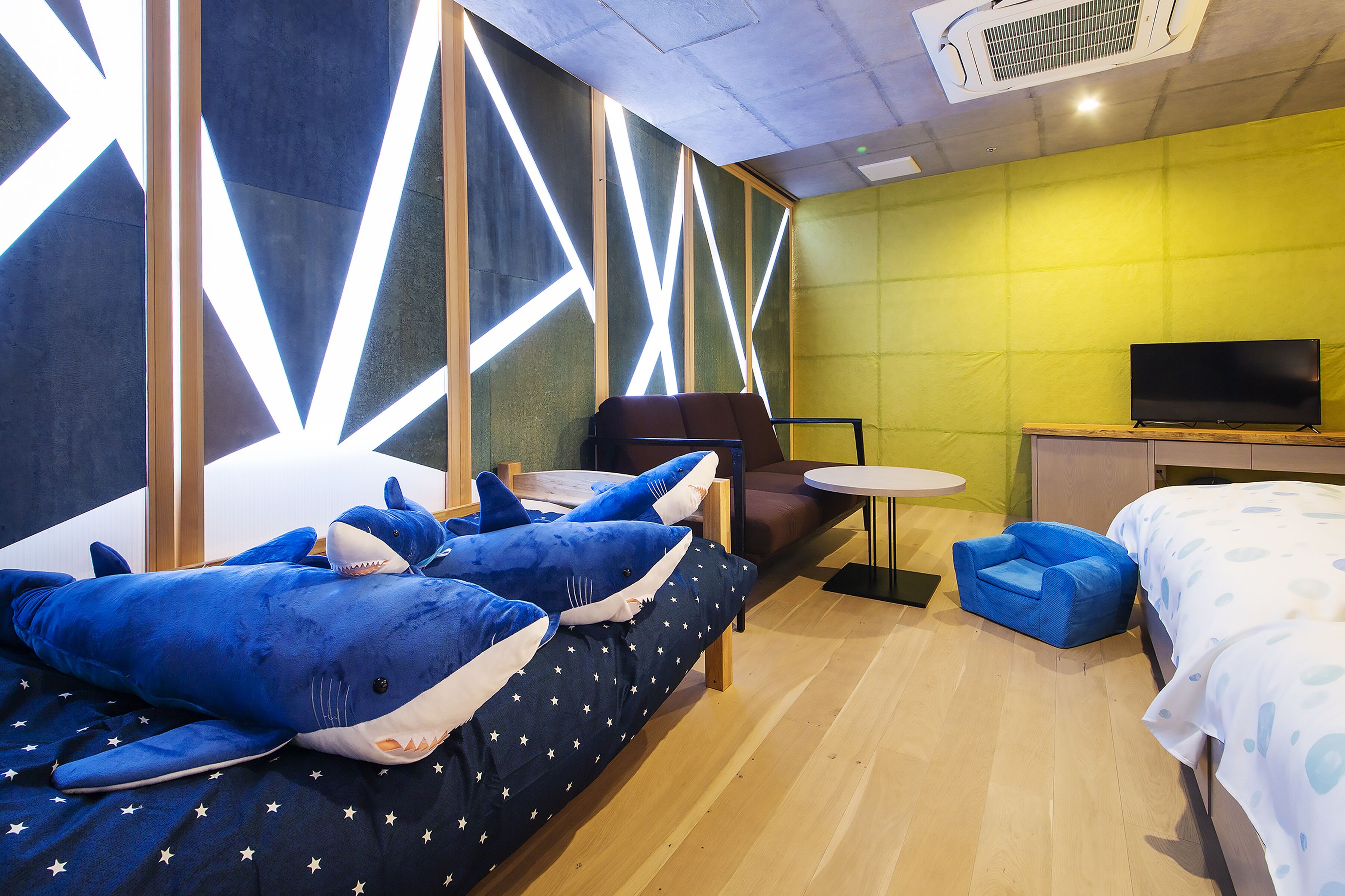 Una habitación de hotel moderna con una cama, un sofá con almohadas con temática de tiburón, un televisor y una mesita.