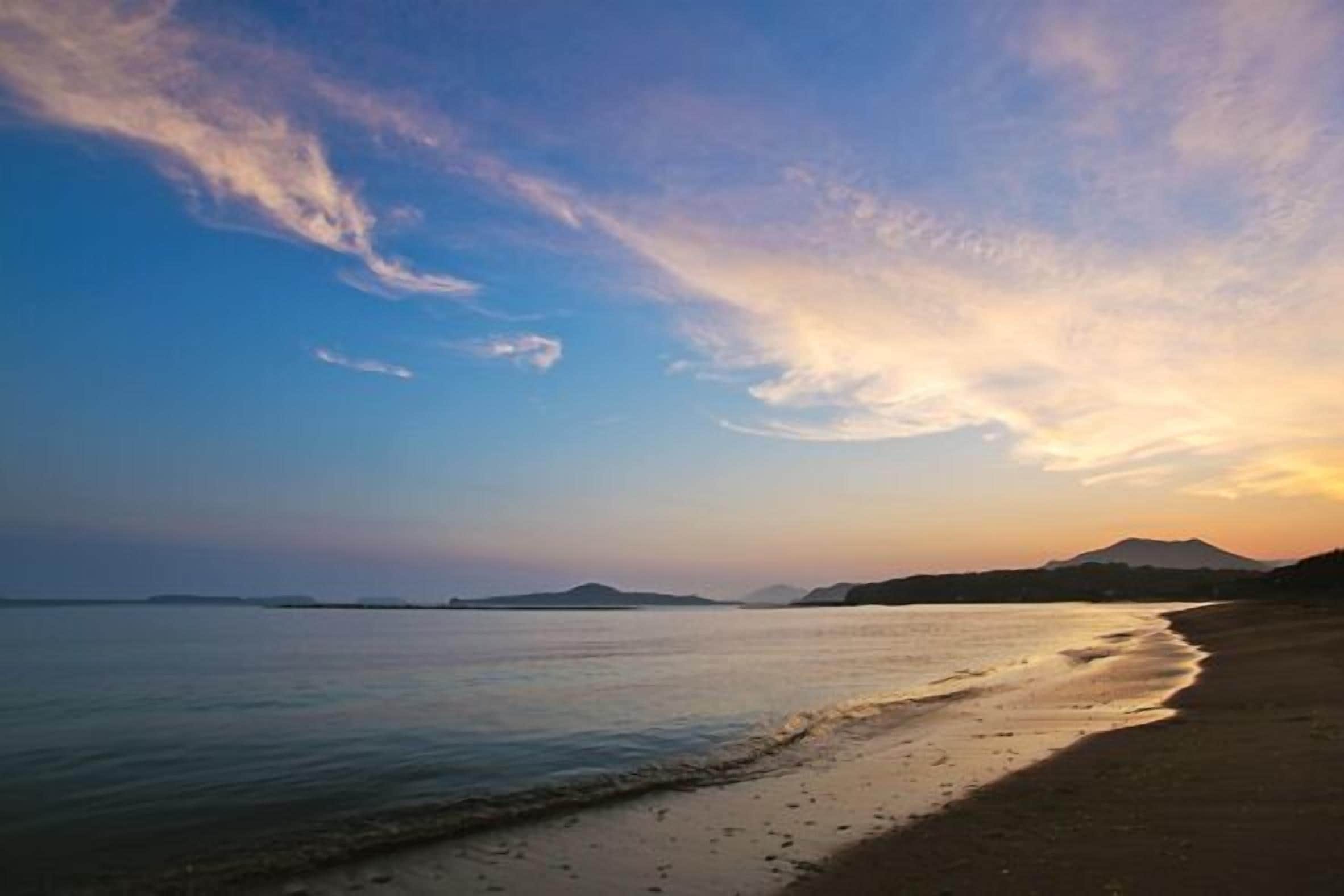 A beach with calm waters and distant mountains under a clear sky.