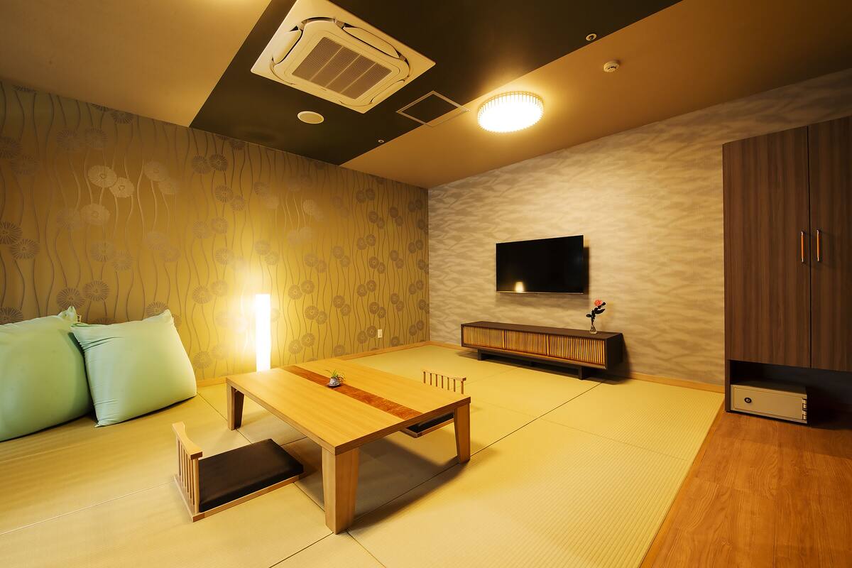 Ein Zimmer mit Tatami-Boden, einem niedrigen Holztisch, einer Couch mit Kissen, einem an der Wand befestigten Fernseher und einem Schrank mit Schubladen.
