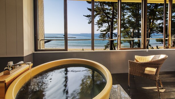 A hot tub with a view of the ocean and trees, a wicker chair, and a wooden side table with a bottle. - Resort Hotel Mihagi (Hagi)