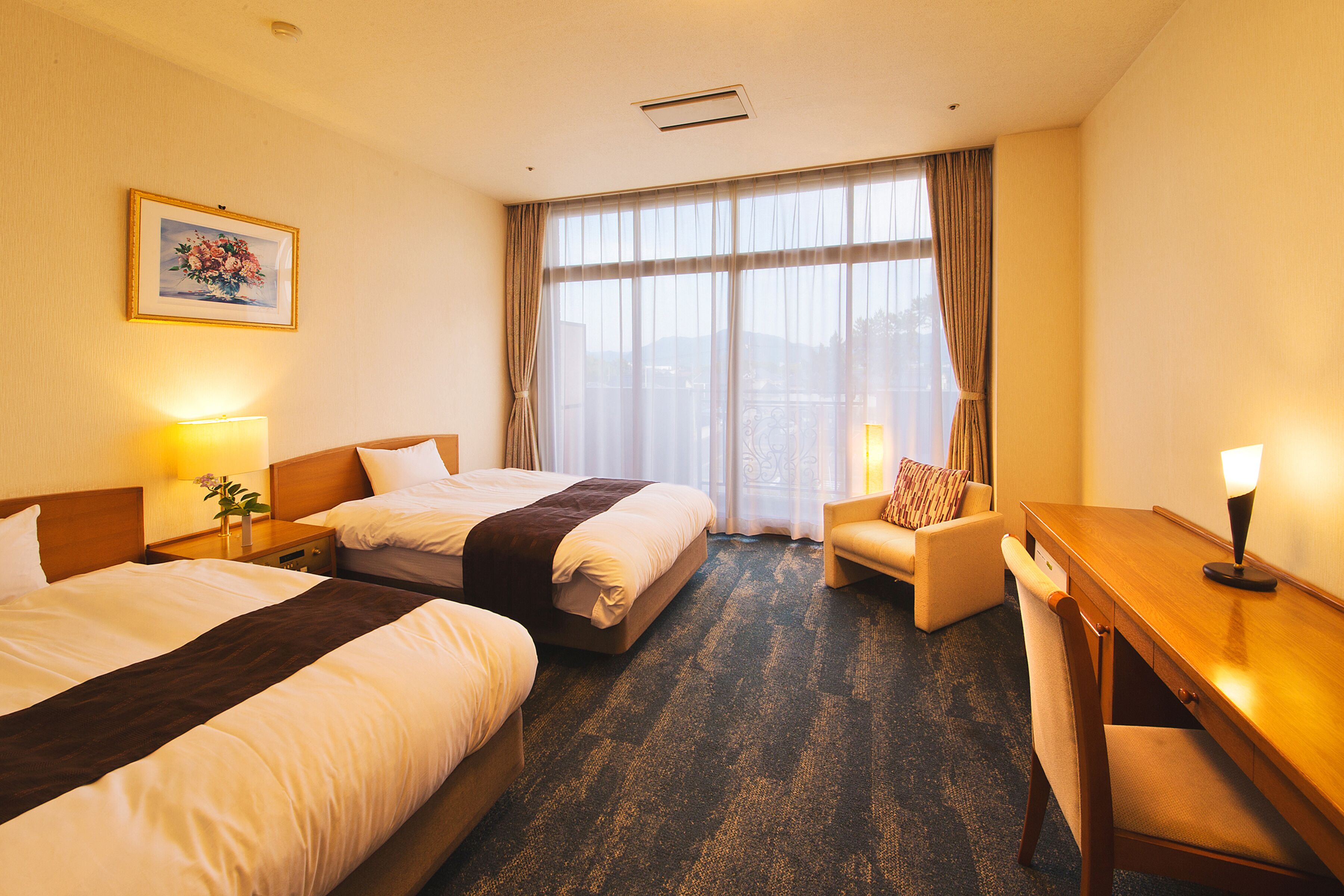 A hotel room with two beds, a desk, a chair, and a large window with curtains.