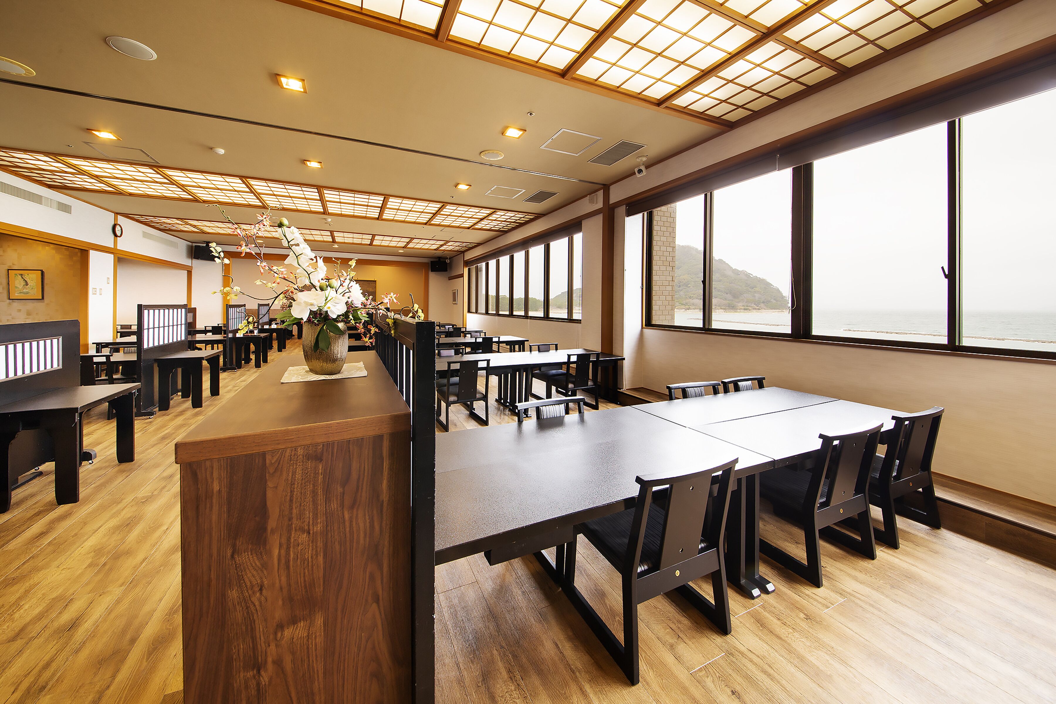 A spacious dining area with wooden tables and chairs, large windows offering a view of the sea, and a traditional ceiling design.