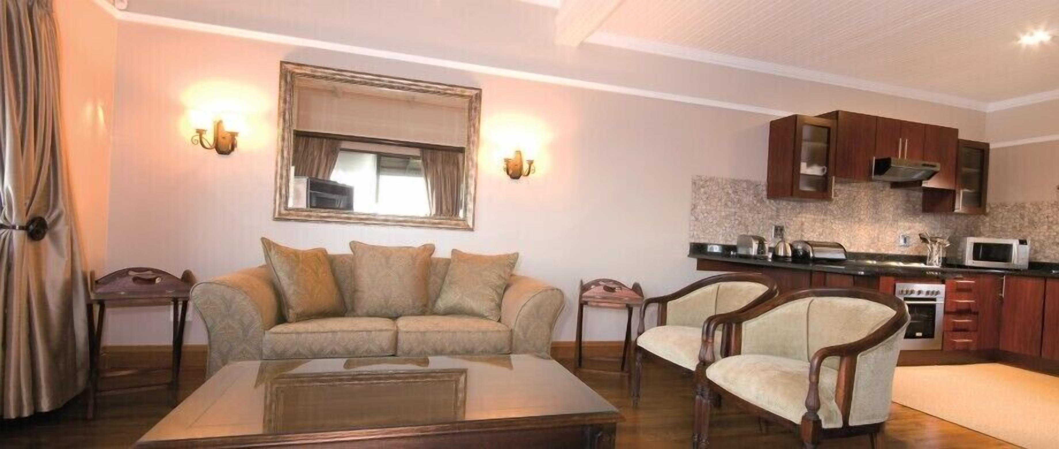 A living room with a sofa, two armchairs, a coffee table, a kitchen with wooden cabinets, a microwave, and a stone backsplash.